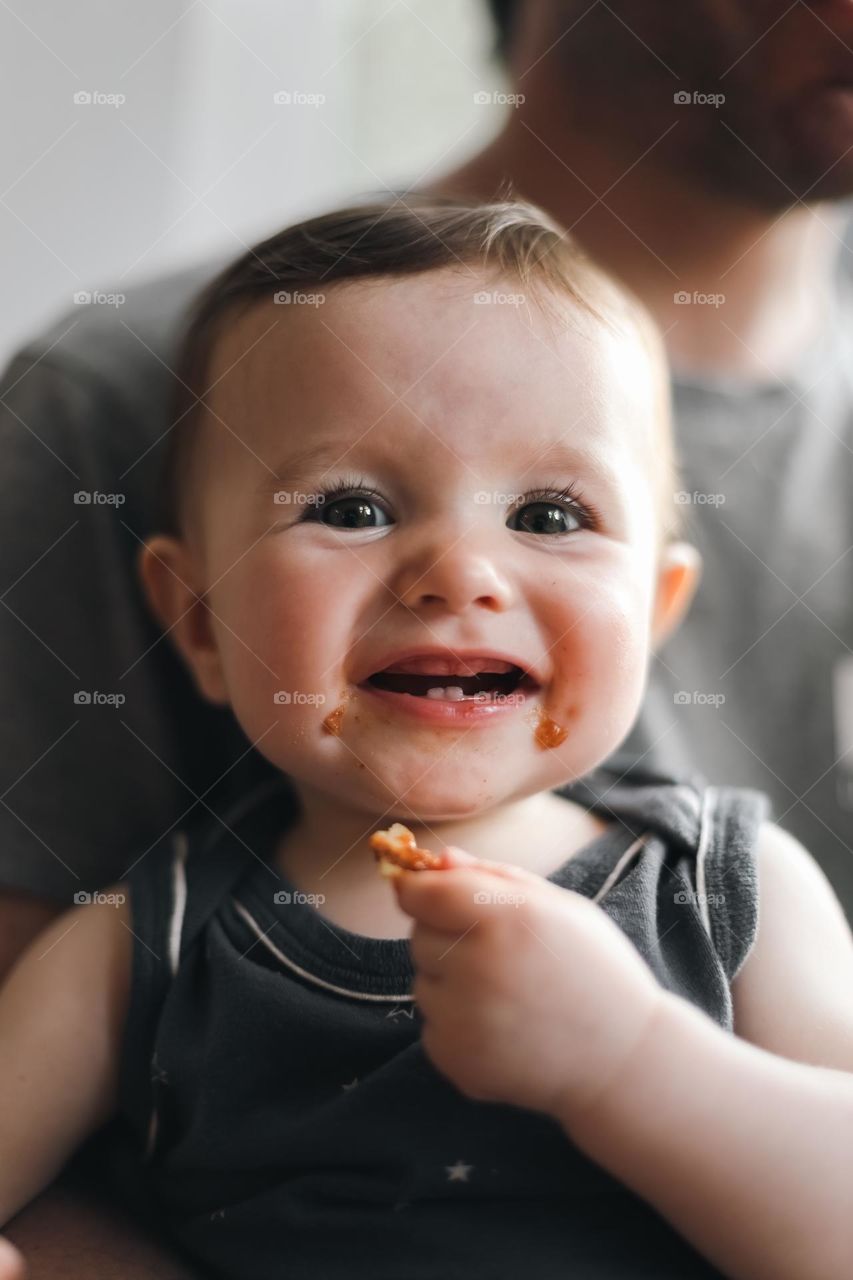 Little caucasian girl with a cute smile, four teeth and a dirty mouth eats a slice of homemade pizza while sitting in her dad's arms at a round table in the kitchen, side view, wow plan.Happy childhood concept, at zoom, dads.