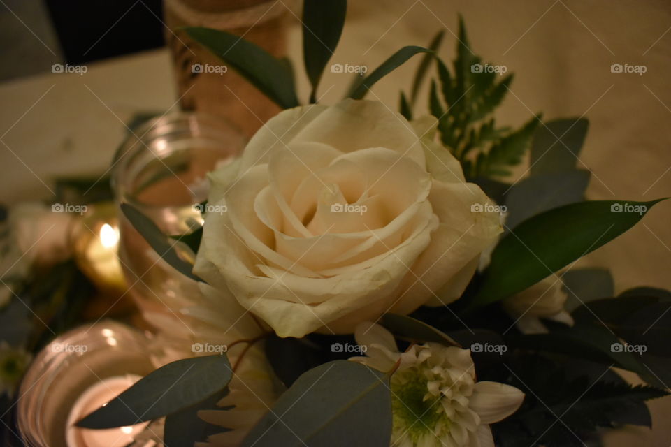 A bouquet featuring an open off white rose, white flowers and rustic greens is back lit by romantic candle light as part of a wedding center piece. 