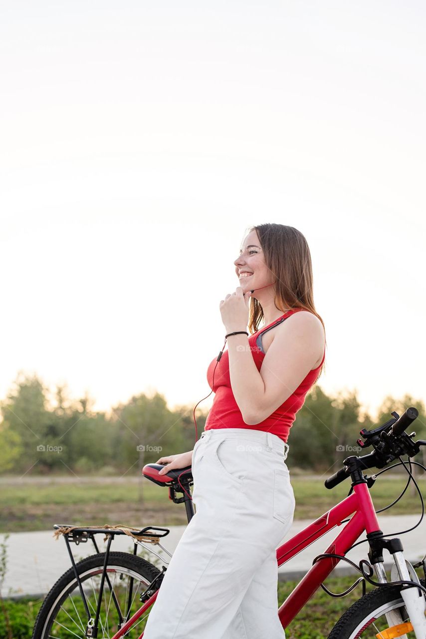 young smiling woman in red shirt and white pants listening to the music standing near her bicycle