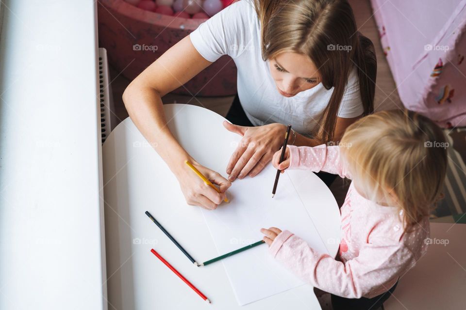 Little girl toddler with her mother drawing with colored pencils on table in children's room at home, view from top