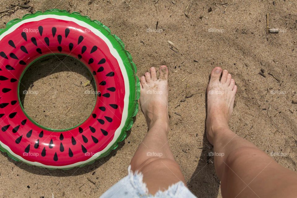 bare legs of a young girl stand on a sandy beach with an inflatable circle in the form of a watermelon