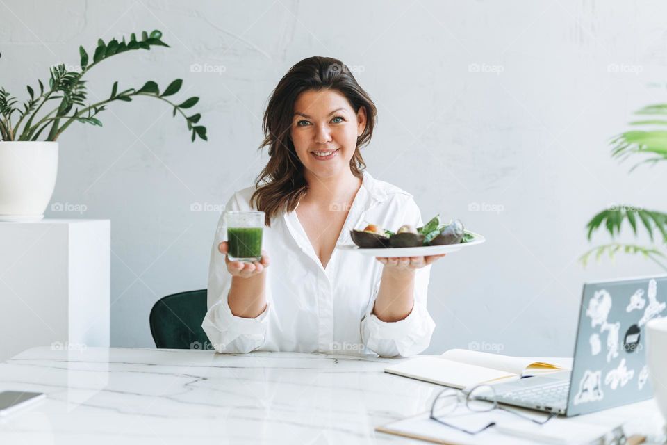 Young smiling brunette woman nutritionist doctor plus size in white shirt with green healthy food at modern bright office room. Doctor communicates with patient online