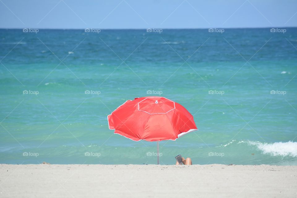 Female reading an electronic book under a red beach umbrella at the seashore with tropical blue ocean in backgrounds and white sand in foreground.