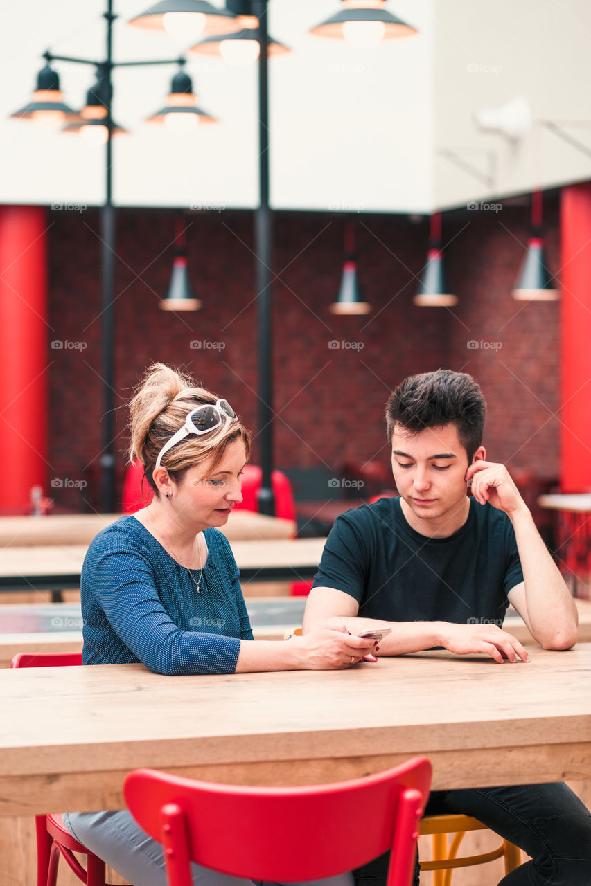 Woman and young man talking together and using mobile phones sitting by a table in cafe