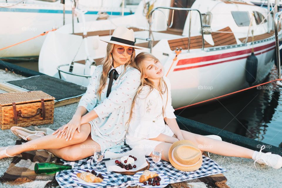 Two beautiful blonde girls friends mother and daughter in straw hats having picnic on yacht pier