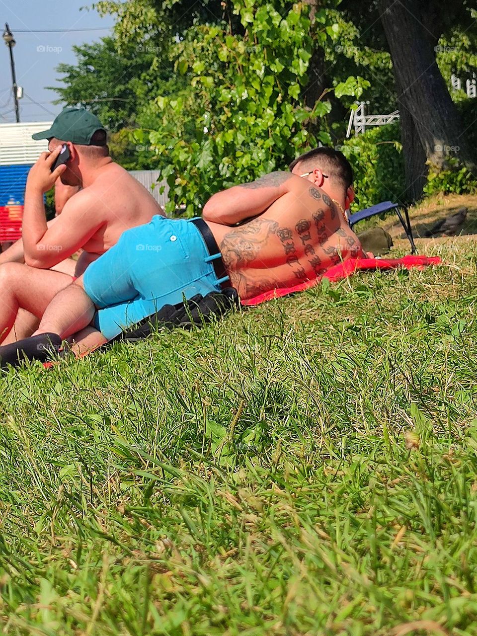Hot summer day. On the bank of the pond are two men who are sunbathing. One man is talking on the phone, the second man is lying on his side. Aesthetic tattoos on the back and on the arm of a man