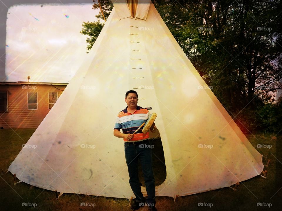 native man stands with his drum by a teepee.