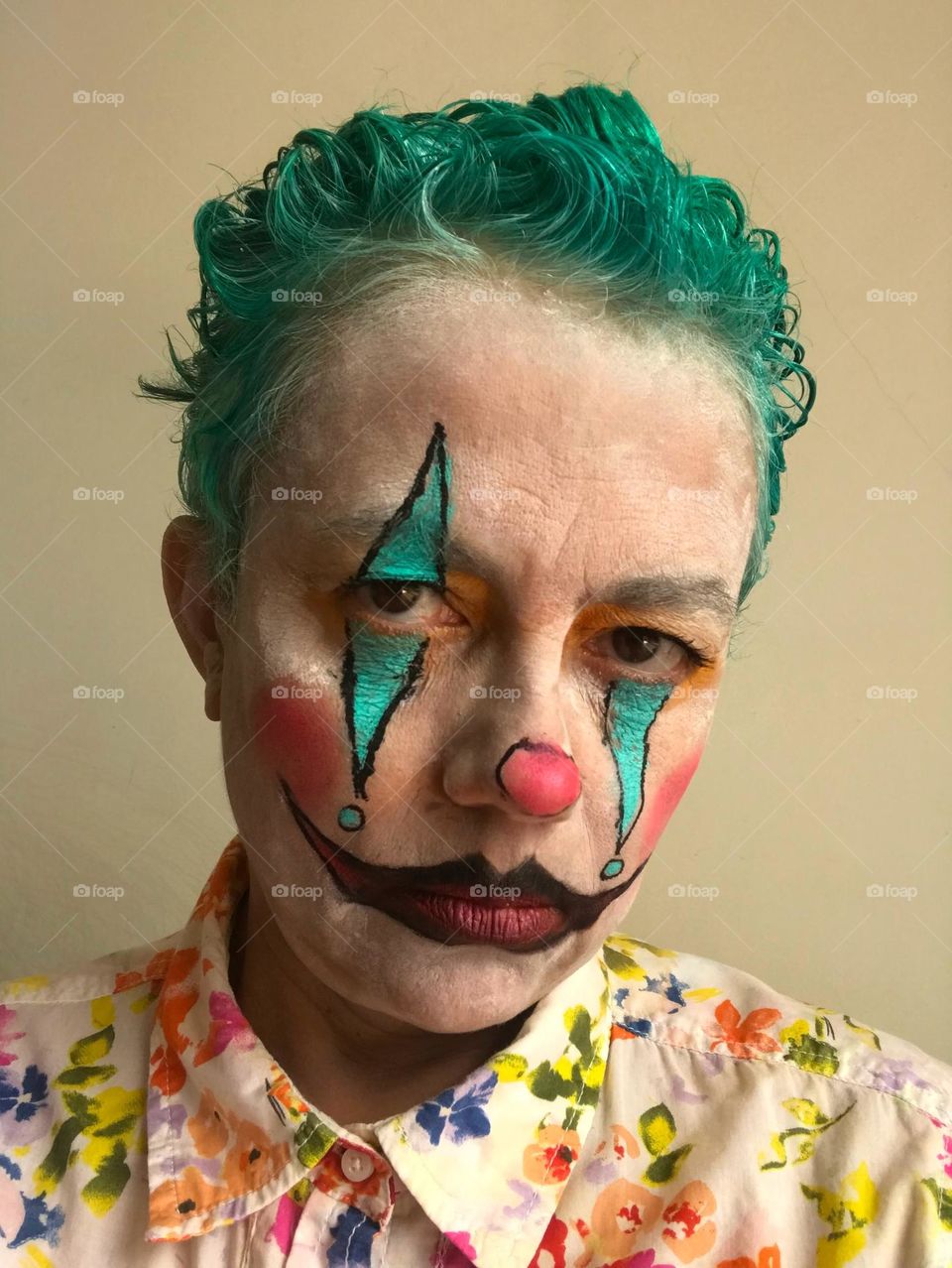 A green-haired joker stares sadly at the audience despite the flowered colorful shirt and the big fake smile painted over the mouth that insists on not smiling.