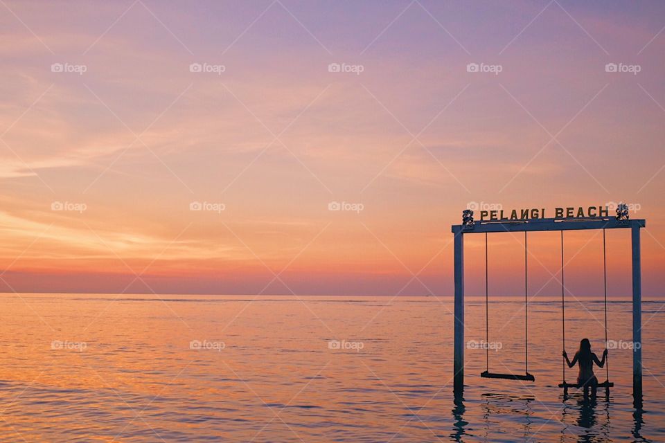 A woman sitting on swings in the middle of the ocean during sunset