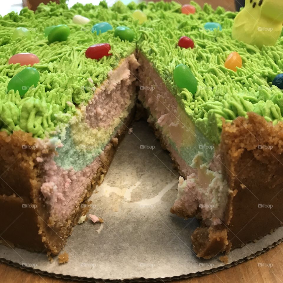 Multicolored Easter cheesecake with slice missing, decorated with green whipped cream grass, jelly beans, and a marshmallow peep. 