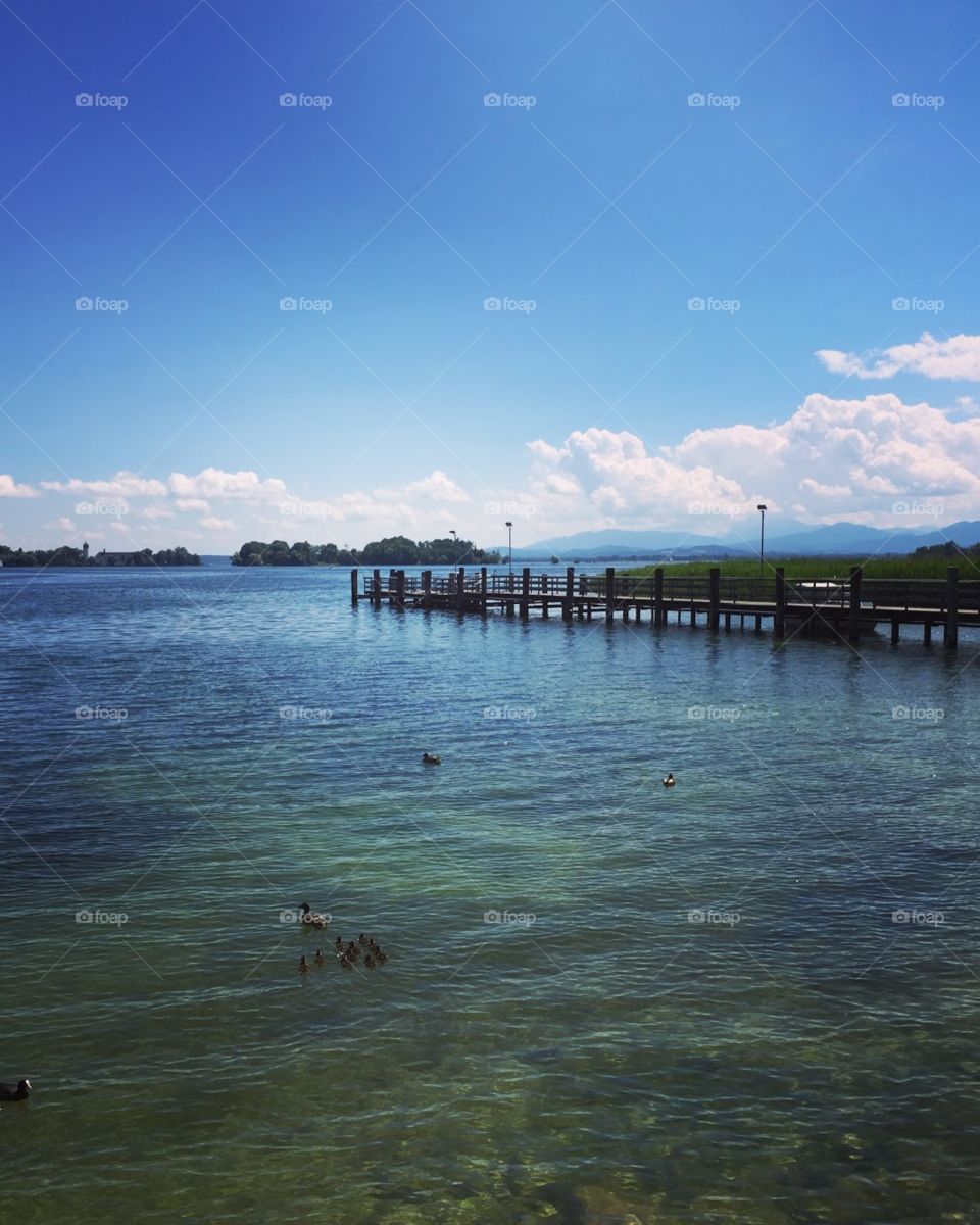 there is a long wooden bridge on the water, the water is very clear, visible on the bottom , there are rows of trees, a lot of mountains in the dítance , blue sky, white clouds