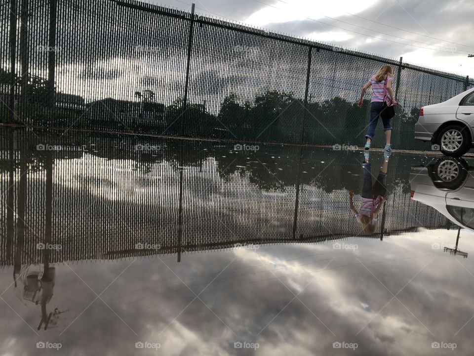 Urban fence and cloudy sky reflected in puddle as girl with blue shoes walks to silver car 