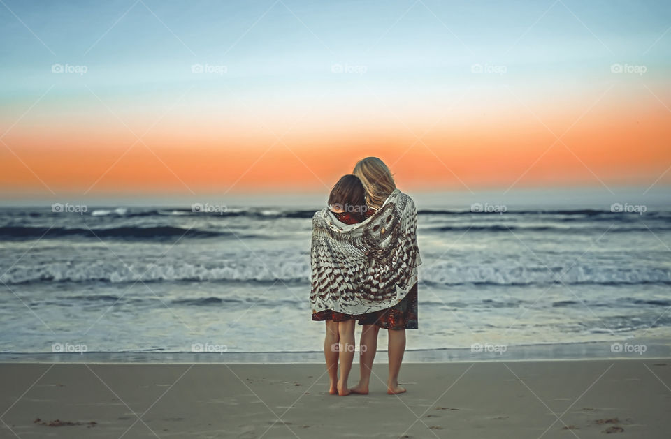 Two girls staying on the beach and looking at the ocean. Sisters or friends stay together covered with one scarf