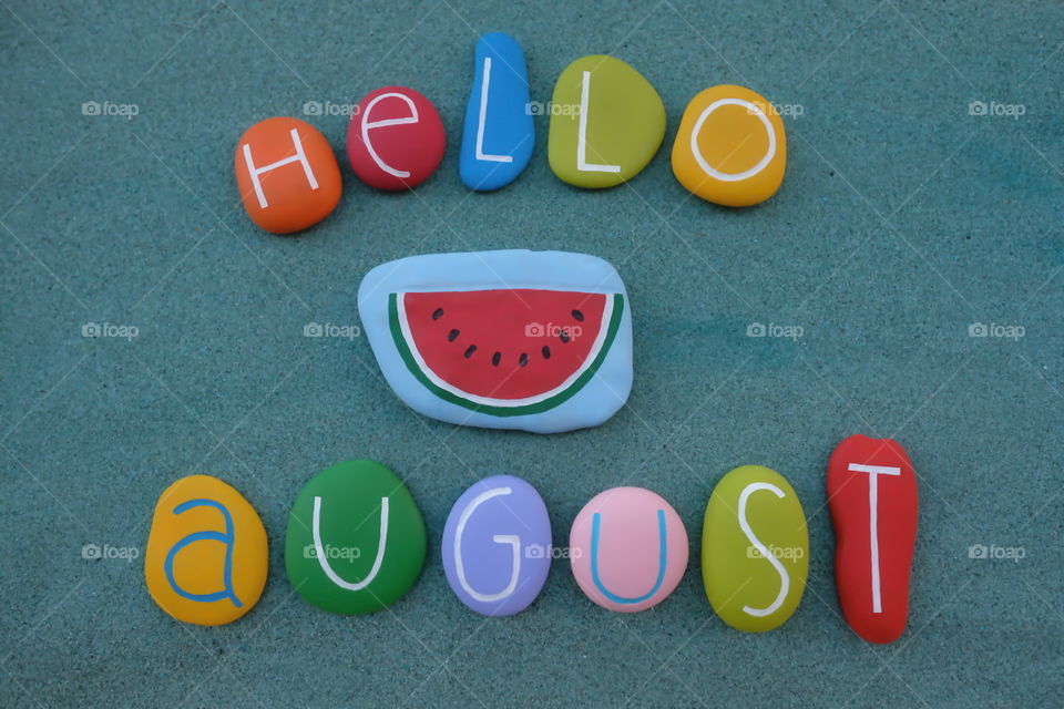 Hello August, summer season month celebrated with a colorful composition of stone letters over green sand 