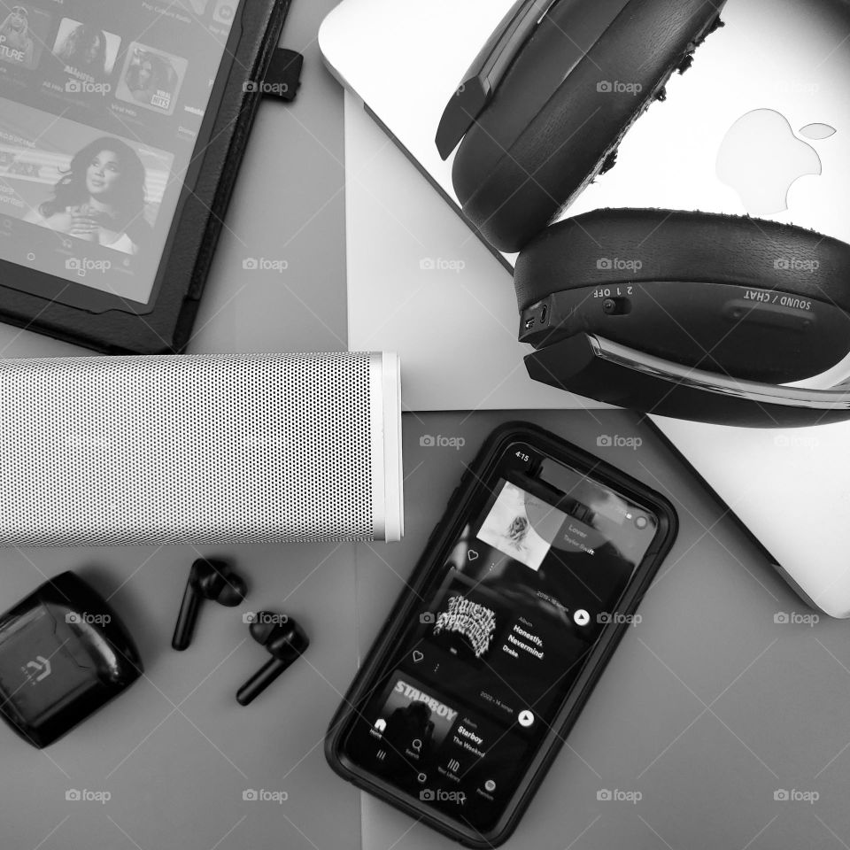 Electronic music listening devices black and white flatlay. Tablet with Amazon music, MacBook air, Bluetooth headset, Bluetooth ear buds, mobile phone, and portable bluetooth speaker.