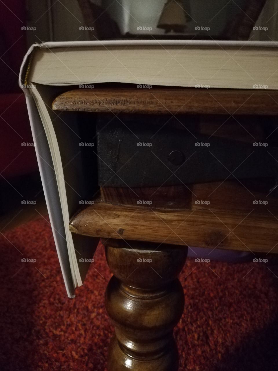An open book on the corner of a brown wooden table. Some pages hanging over the edge. A hammered black iron decoration with a nail on the side of the table.