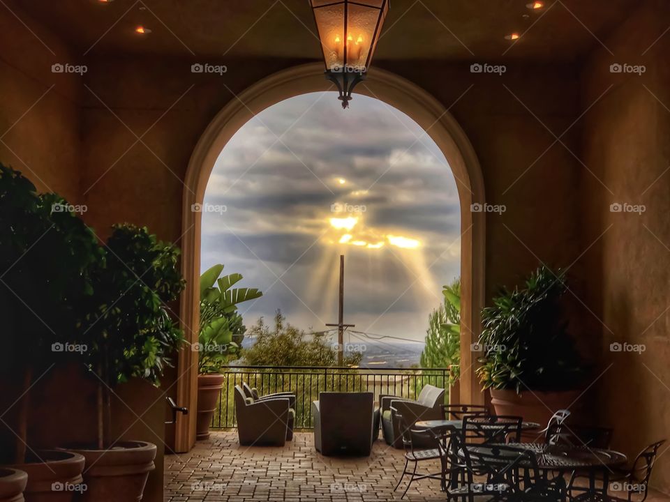 Peering out from a balcony with the sun shining through the clouds with chairs placed in view and trees in the outer frame. A cross in center frame