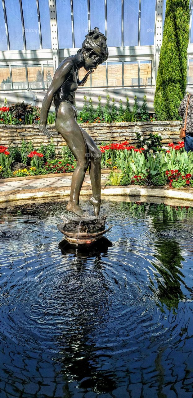Statue of a young nude in a reflecting pool with Spring of red Tulips in the background while Sunny skies reflecting into the pool.