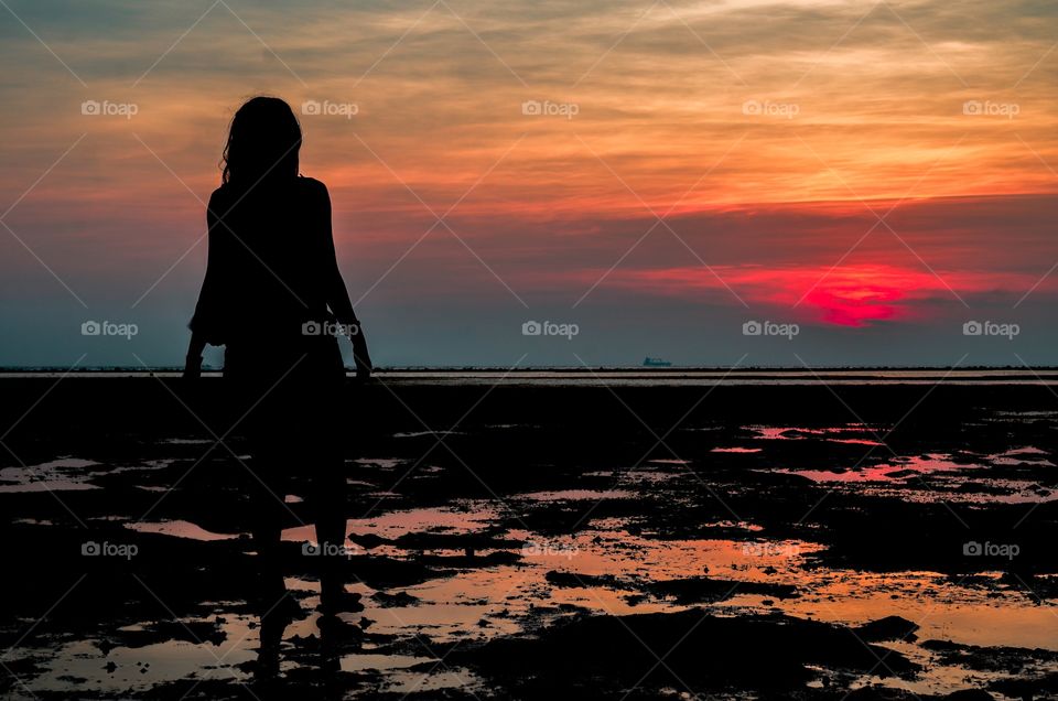 A young woman walking by the beach while the sun is setting in the background.