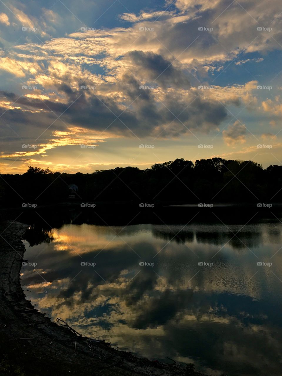 Clouds and sunset illuminate the skyline over river and silhouetted trees, with reflection 