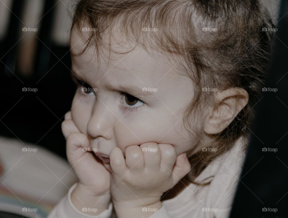 Portrait of a beautiful little Caucasian girl with an angry emotion on her face resting her cheeks with both hands, close-up side view.
