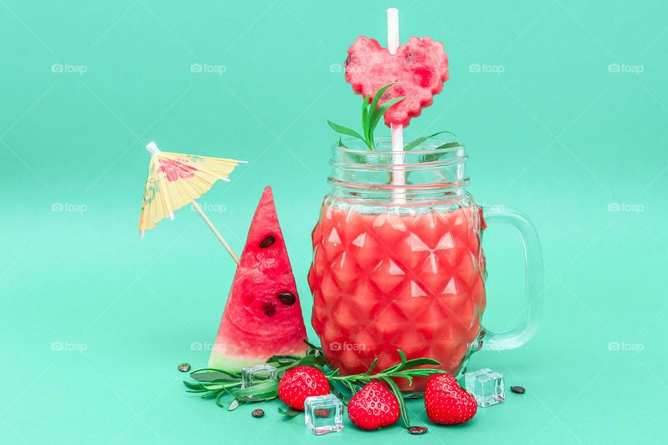 Fresh watermelon and strawberry drink in faceted jar with metal lid, handle, straws, triangle shaped watermelon slices and heart, ice cubes, rosemary branches, strawberries and skewer with paper umbrella on green background, closeup side view.