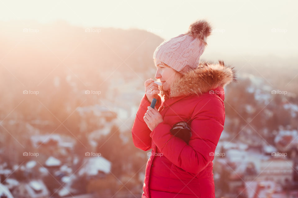 Woman applying lip balsam while walk on a wintery day. Wearing red coat and cap. Town in the background