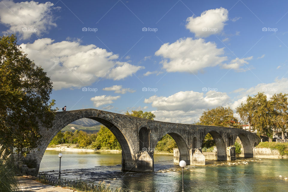 Stone bridge over a river in Greece, shot in a sunny but also a bit cloudy day.