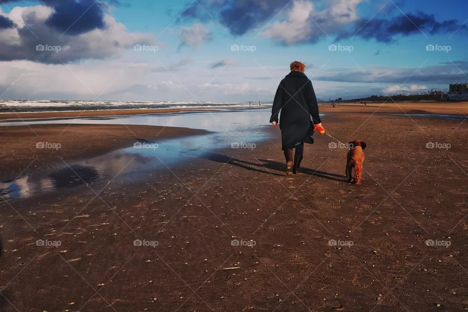 Woman walking on the beach with her dog on a beautiful sunny winter day, blue sky with clouds