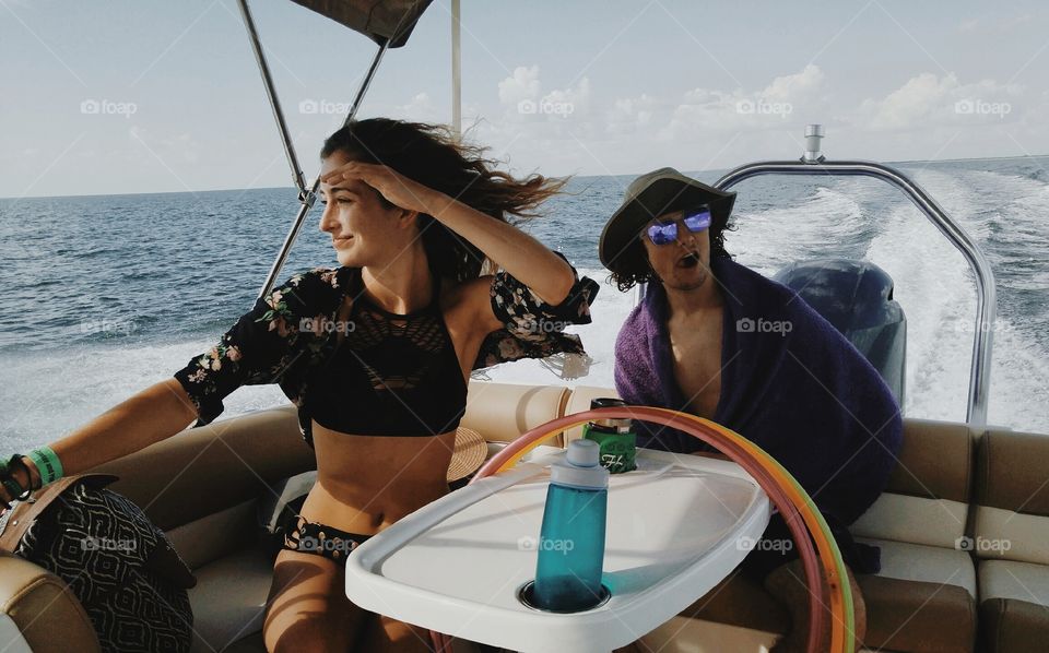 a girl with her hand on her head looking over the water from a boat like a pirate next to her brother who is acting silly sticking his tongue out wearing a hat and towel