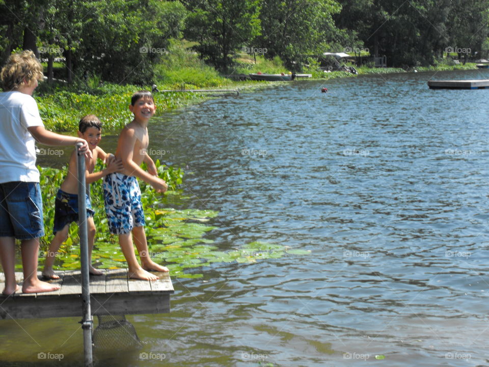 Three boys standing on the end of a pier. One boy will push another off and into the water.