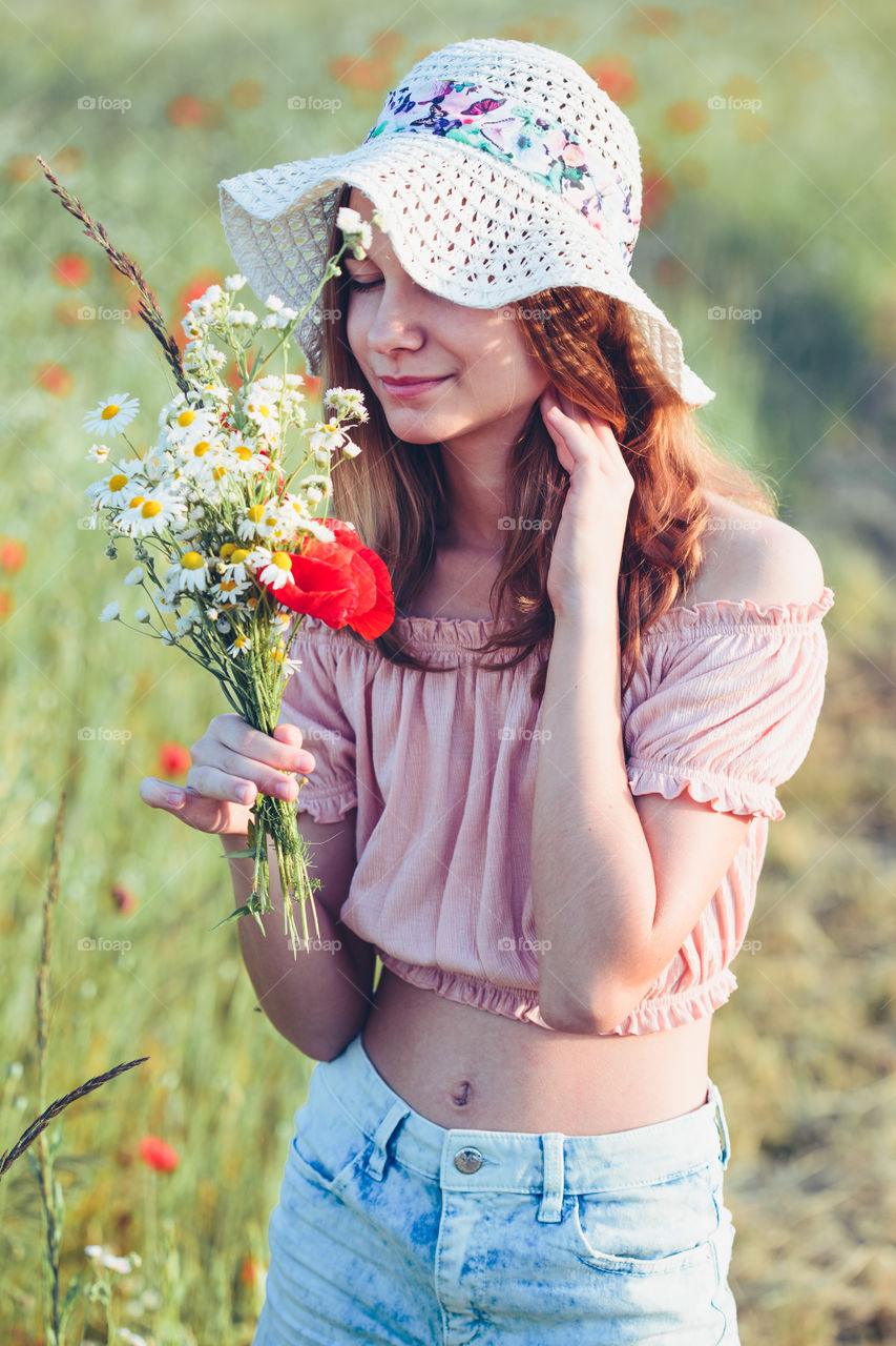 Beautieful young girl in the field of wild flowers. Teenage girl picking the spring flowers in the meadow, holding bouquet of flowers. She wearing hat and summer clothes. Spending time close to nature