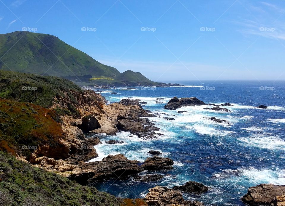 Bright and brilliant California coast with blue ocean and rolling green mountains.
