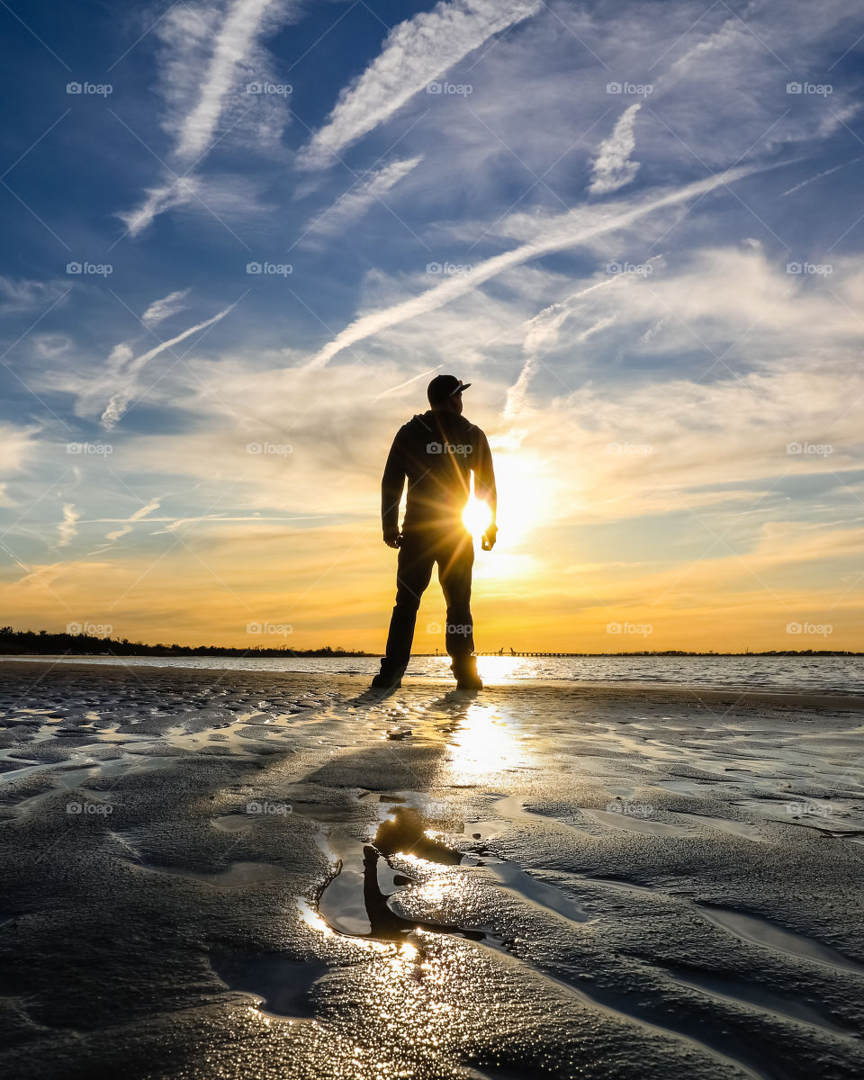Silhouette of a young man on the beach with sun rays shining across his body as the sun sets over the water