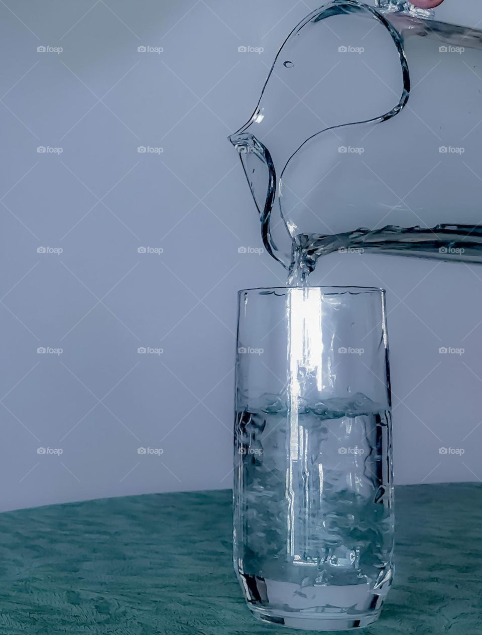Pouring pure water from a clear glass pitcher into a clear water glass on a dark green covered table. 