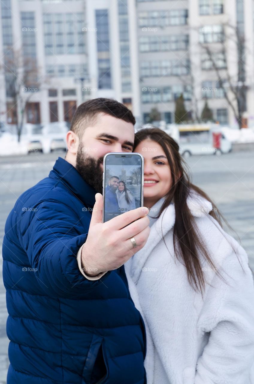 Portrait of young happy couple, bearded man and brunette woman woman relaxing in winter city. Laptop. Phone. Technology. Selfie.  Social media.  Video call