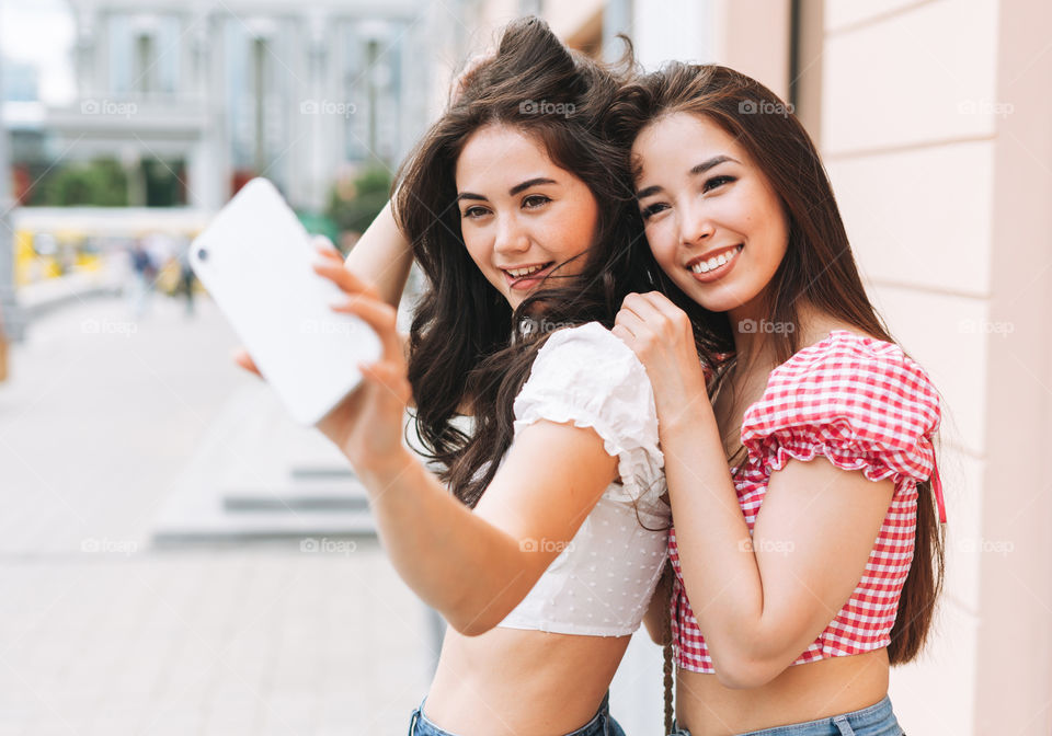 Happy smiling beautiful brunette young women friends in summer clothes taking selfie on mobile phone at the summer city street