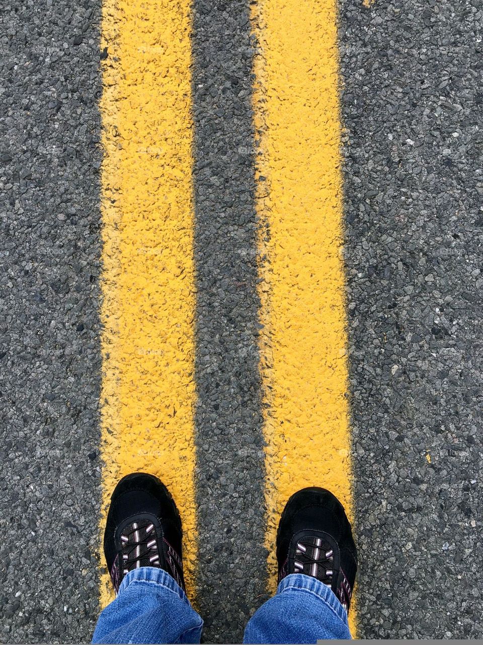 Standing on double yellow lines in street. I'm trying to decide which way to go, left or right? Black sneaker tips, time to walk. Double yellow lines in middle of the road. Black asphalt & two yellow lines means no passing.