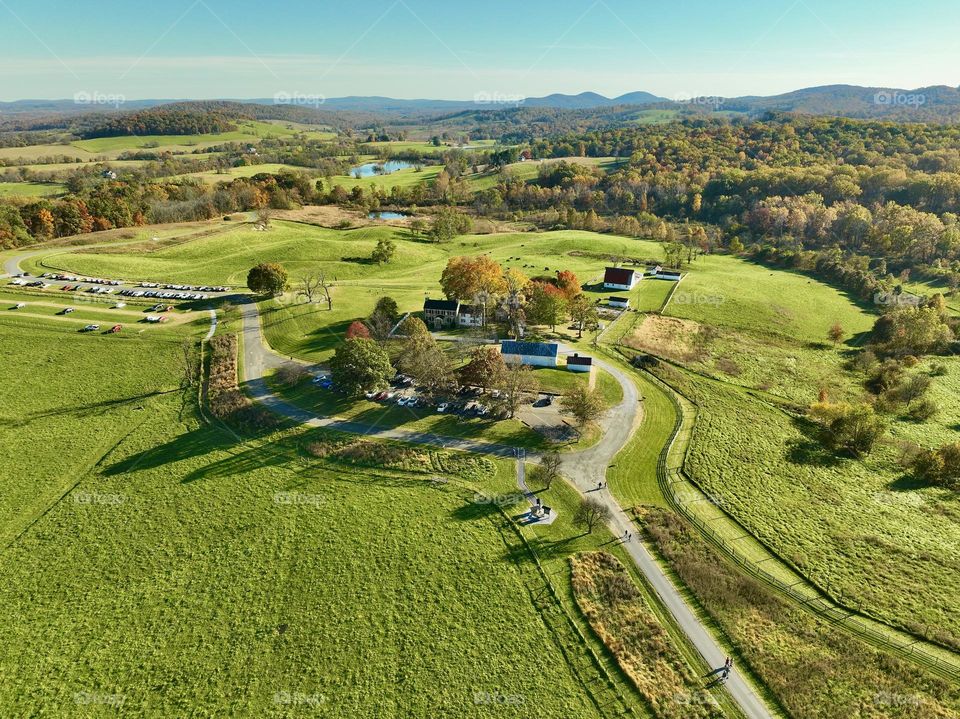 Countryside Harmony: An aerial view of a scenic farmstead surrounded by lush green fields, winding roads, and distant rolling hills beneath a crisp autumn sky.