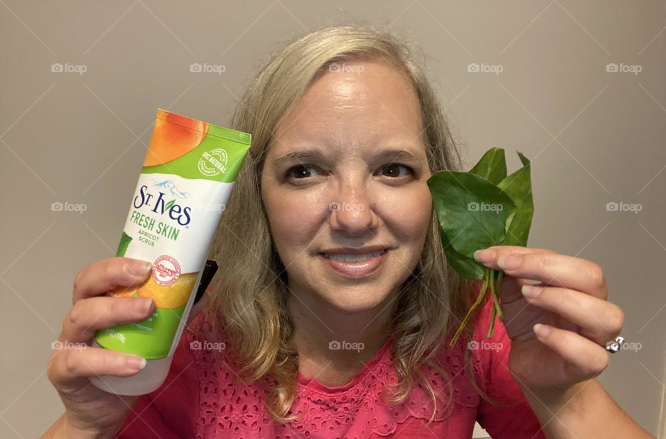 A woman (myself)holding St. Ives Fresh Skin Apricot Scrub in one hand and green plant leaves in the other. I love this product because it makes skin soft and has natural ingredients. Nature and this scrub go hand in hand. 