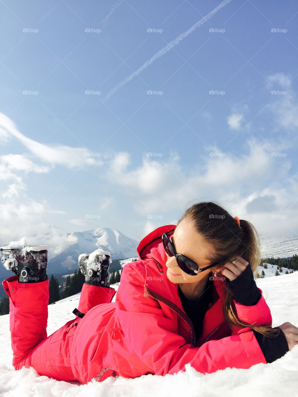 Smiling woman with sunglasses dressed in pink ski costume laying on snow with snow mountains in the background