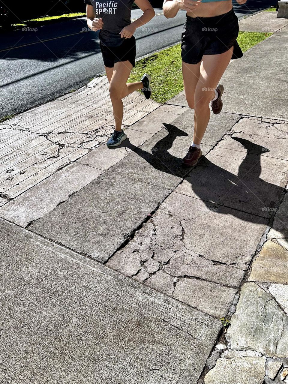 Two female runners in black shorts running side by side with matching strides down a cracked cement sidewalk next to a road