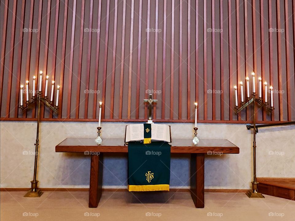An alter setting with a beautiful table with candles on the side, a large open bible on a stand, and green and yellow paraments draped from the table. 