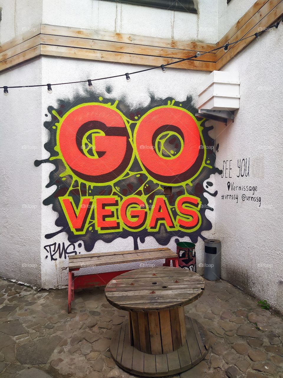 Corner of the white house.  Wooden table and bench.  Graffiti on the wall "GO Vegas"