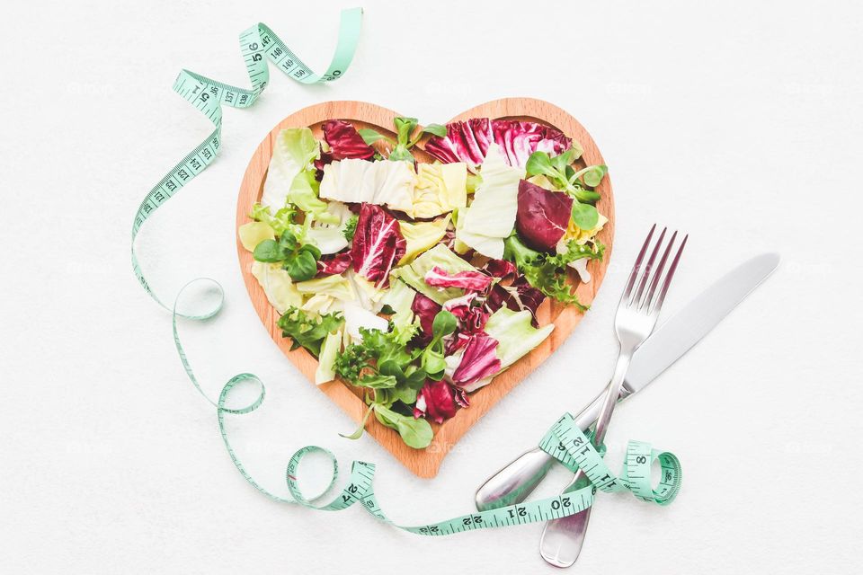 Fresh sliced ​​lettuce in a heart-shaped wooden plate with a fork, knife and measuring tape lie in the center on a white cement background, flat lay close-up. The concept of healthy eating, healthy lifestyle.