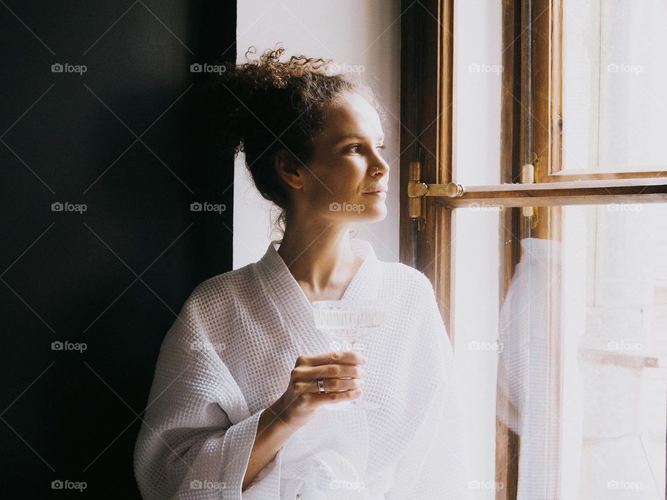 Young beautiful woman with curly hair wearing white bathrobe sits on the window in the bathroom and drinks champagne, portrait of woman, looking at window, lifestyle, at home, relax.