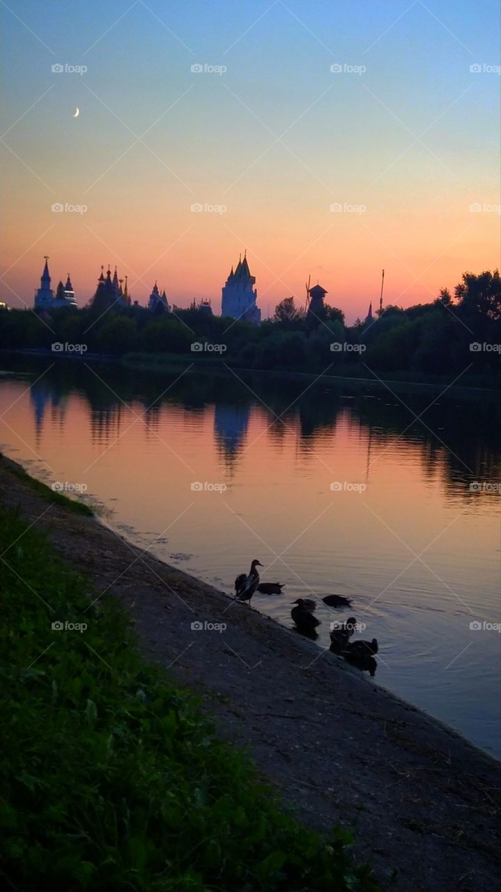 Sunset on the pond.  All shades of pink.  A bunch of ducks on the shore.  In the distance, the domes of the towers among the trees.  There is a crescent moon in the sky.  All objects are reflected in the water