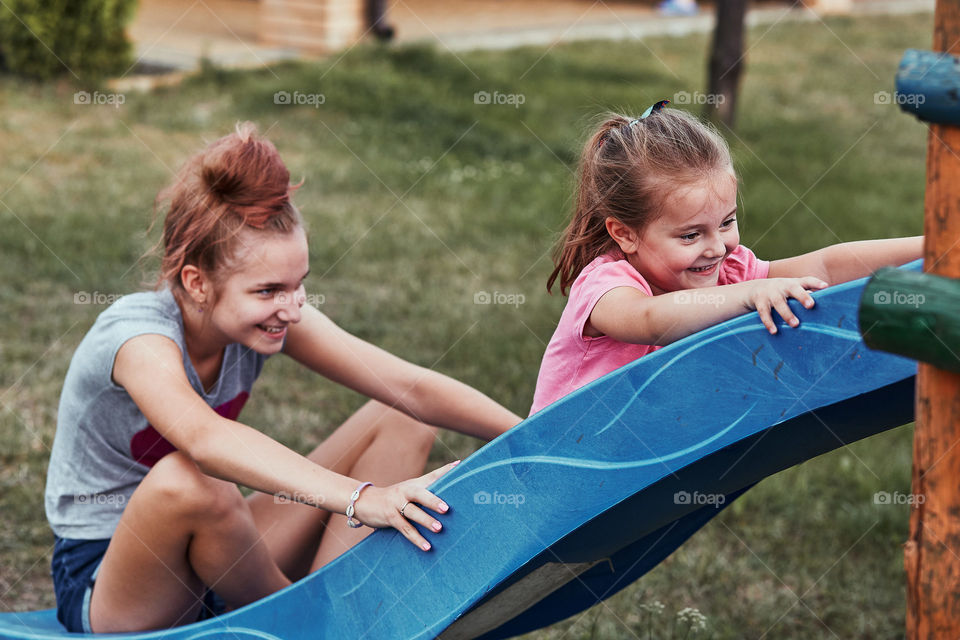 Teenage girl playing with her younger sister in a home playground in a backyard. Happy smiling sisters having fun on a slide together on summer day. Real people, authentic situations