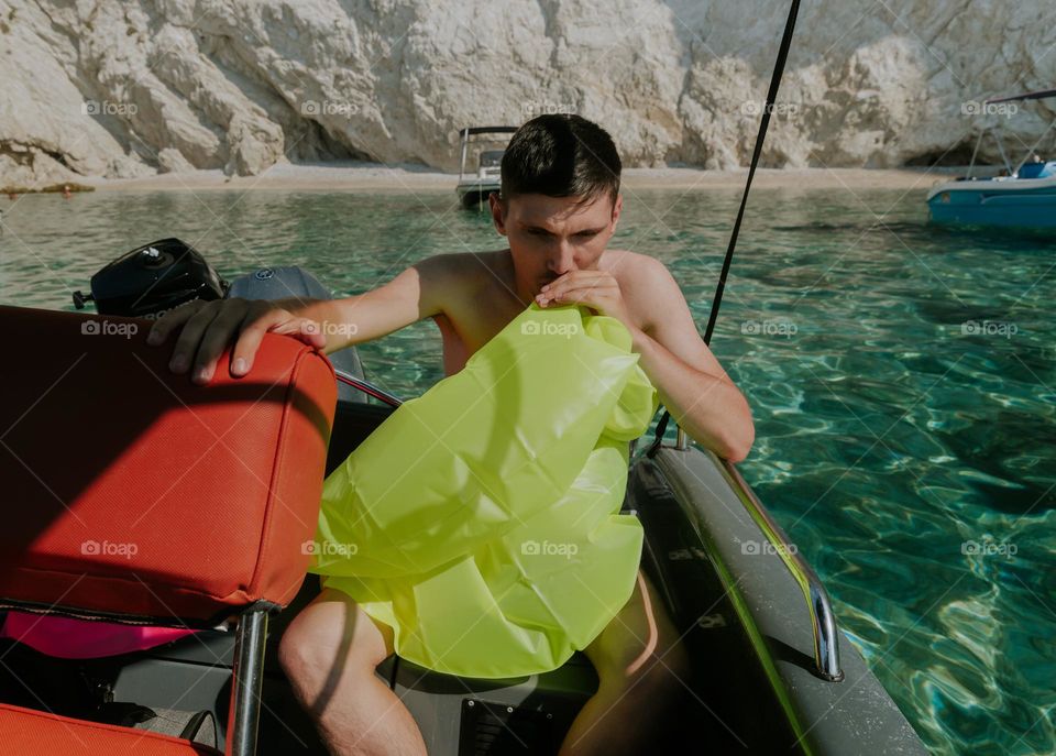 Portrait of a handsome young Caucasian guy inflating a lettuce life preserver sitting in a boat and holding the seat with his hand against the background of rocks in the sea on a sunny summer day, close-up view from the side.