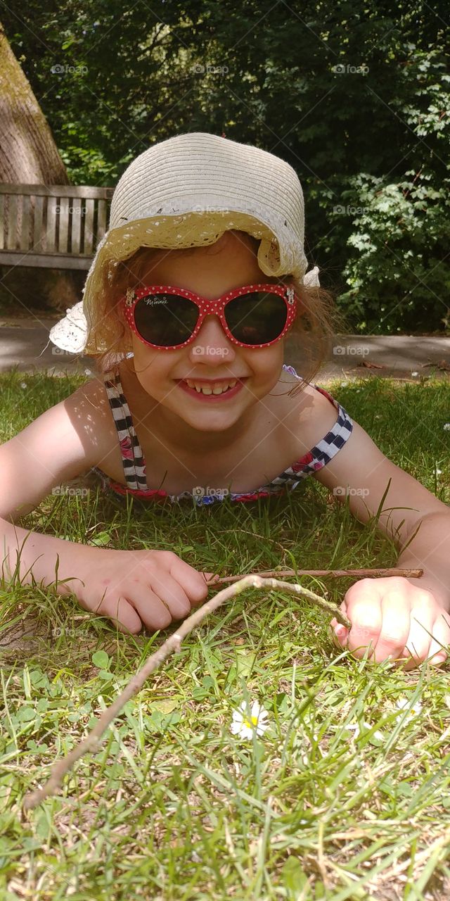 child lying in grass with sun hat and sun glasses smiling at camera.